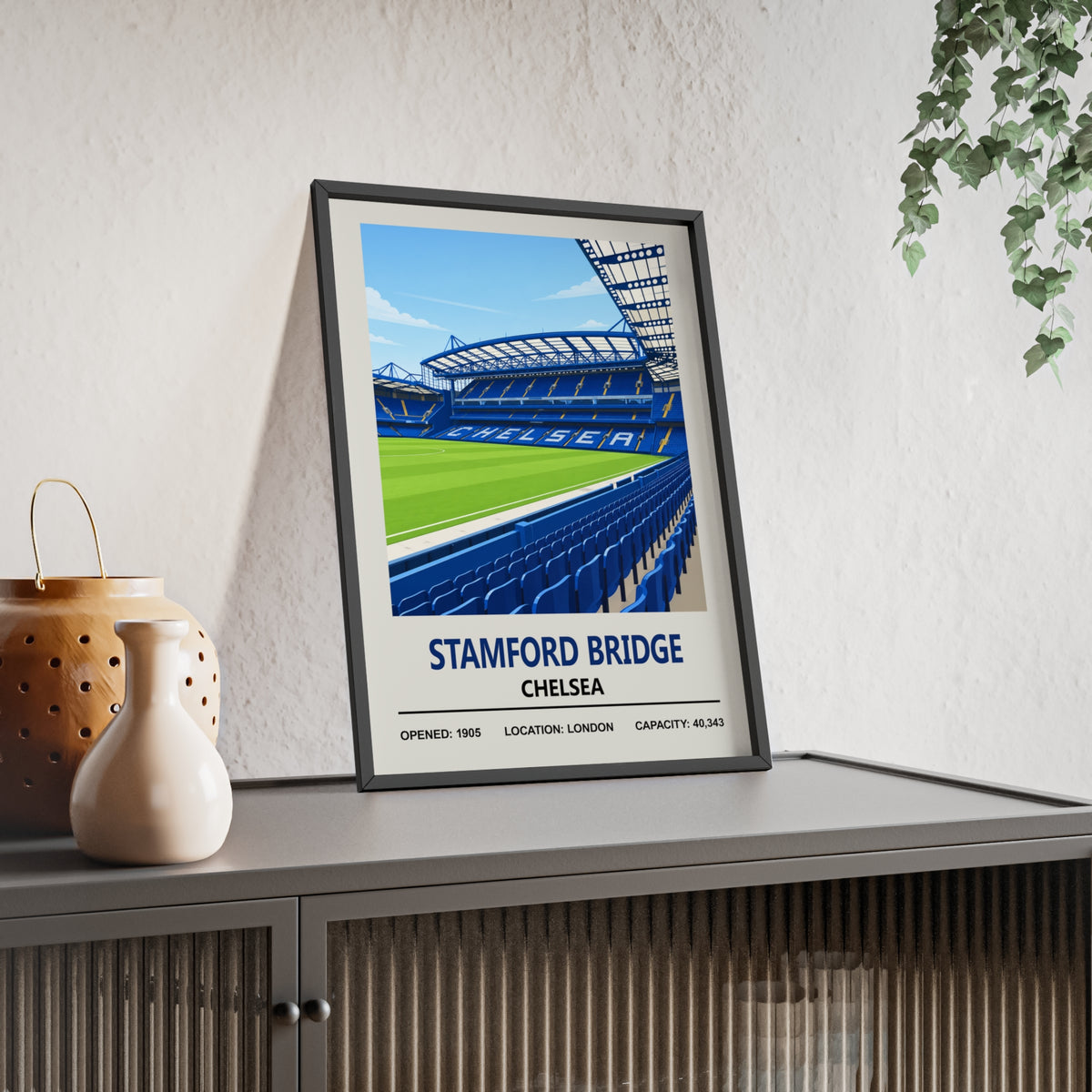 Chelsea Stamford Bridge stadium print in a black frame, leaning on a sideboard beside a vase and trailing plant, showing the blue stand seating, pitch and CHELSEA lettering with opened 1905 and 40,343 capacity