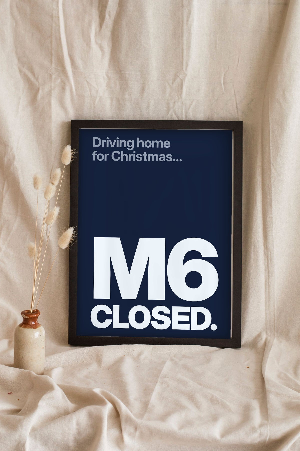 Funny Driving Home for Christmas lyric print in a black frame propped up on linen