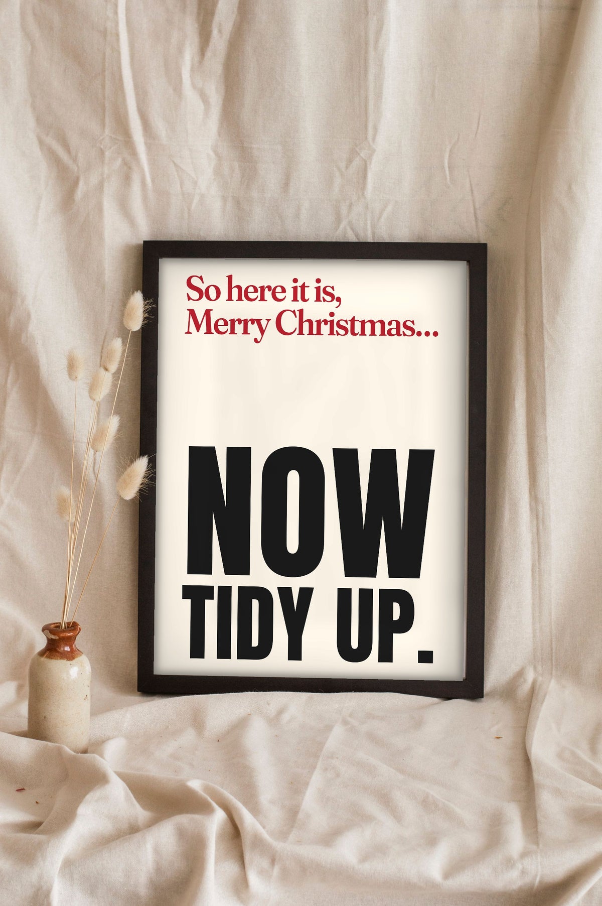 Funny Merry Xmas Everybody lyric print in a black frame propped up on linen