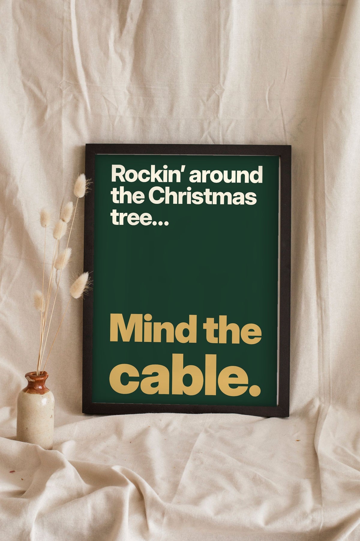 Funny Rockin' Around the Christmas Tree lyric print in a black frame propped up on linen