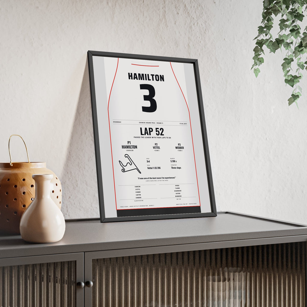Lewis Hamilton 2011 Chinese Grand Prix Win Poster - 3 Race Suit Print framed and leaning on a sideboard