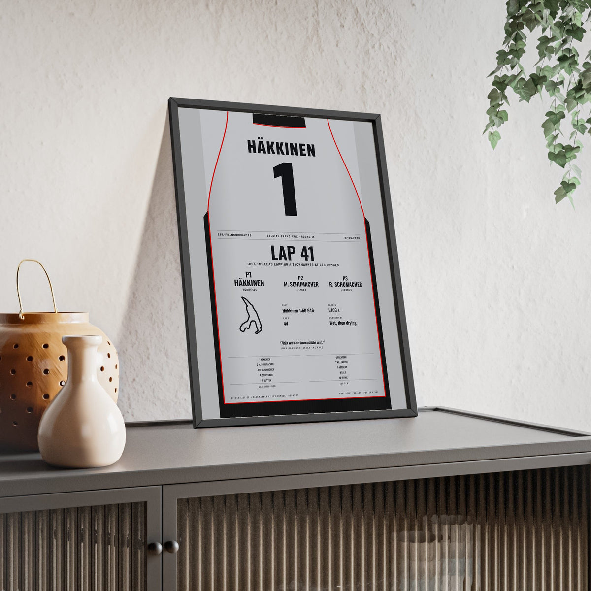 Mika Hakkinen 2000 Belgian Grand Prix Win Poster - 1 Race Suit Print framed and leaning on a sideboard