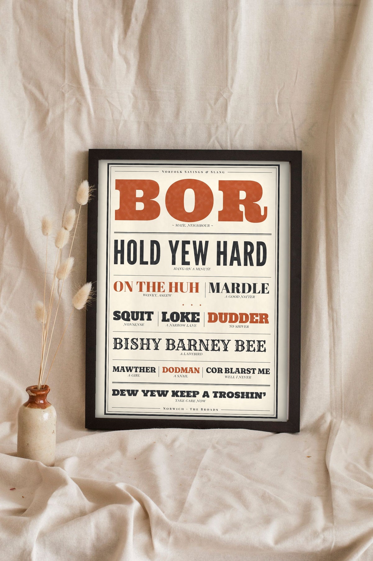 Norfolk Slang Print in a black frame propped up on linen